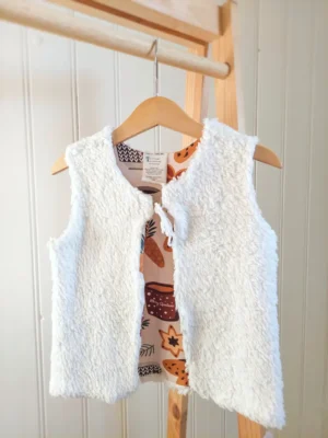 Gilet de Berger réversible motif Ambiance de Noël, face fausse fourrure à l’extérieur, suspendu à un cintre sur un portant en bois.