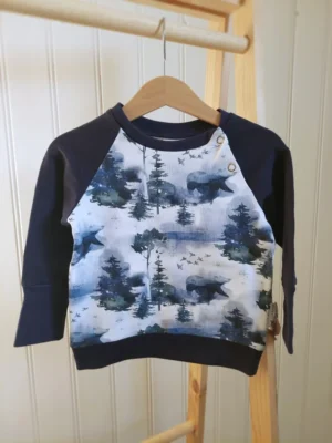 Sweat évolutif taille 12/24 mois, motif Paysage d’Hiver, accroché à un cintre sur un portant en bois.